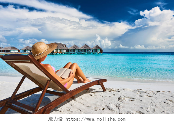 蓝天白云下在海滩边上坐在沙滩椅上j看书的美女读一本书在海滩上的年轻女子
